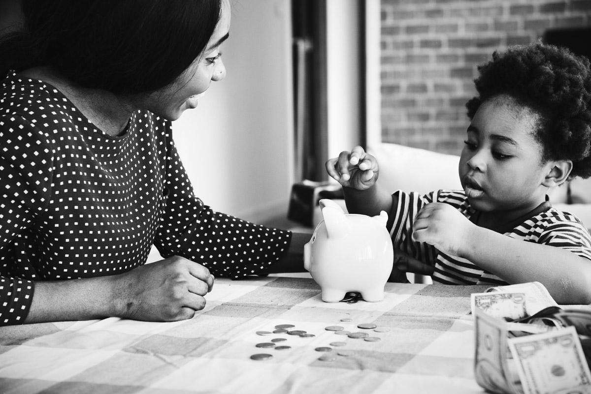 Black and white representation of a small child putting his money in a piggy bank in front of his mother by sitting on the table.